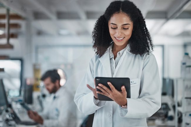 Scientist in a white lab coat using a tablet in a laboratory, with another researcher working in the background.