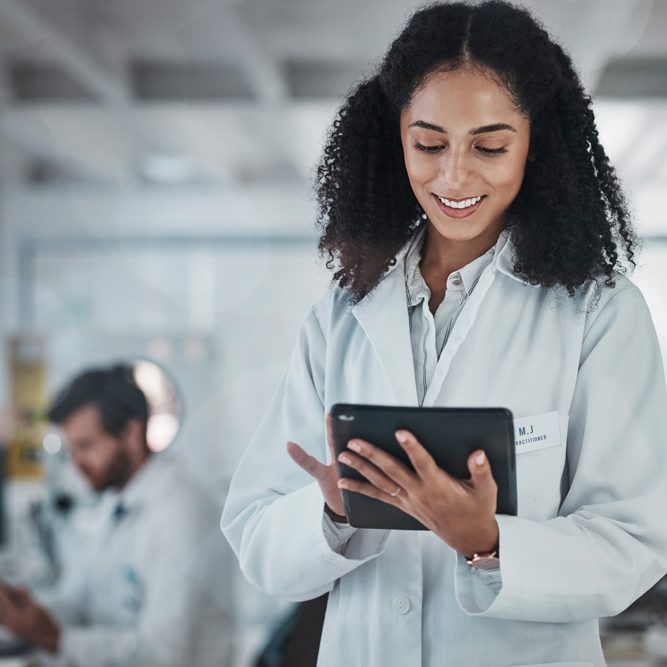 Scientist in a white lab coat using a tablet in a laboratory, with another researcher working in the background.