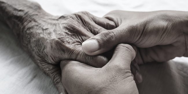 Caring hands grasp the hands of an older person on a white bed in a hospital.