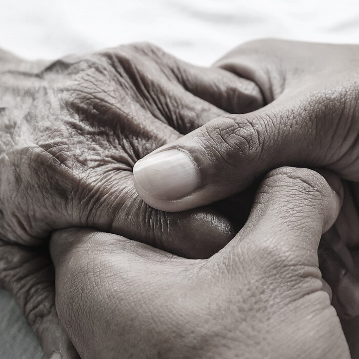 Caring hands grasp the hands of an older person on a white bed in a hospital.