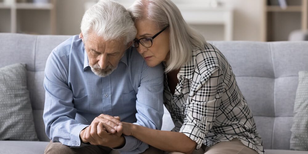 Loving older wife comforting her confused husband, sitting on sofa.