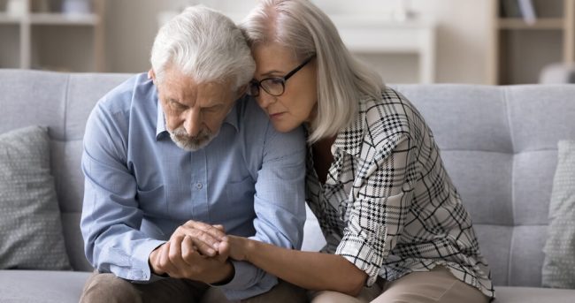 Loving older wife comforting her confused husband, sitting on sofa.