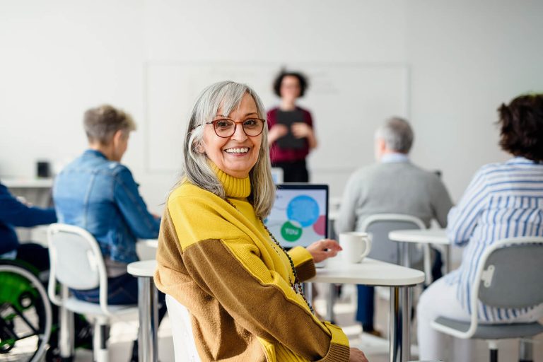 Smiling older woman in a classroom setting using a laptop, with other adults seated at tables and a presenter speaking in the background.