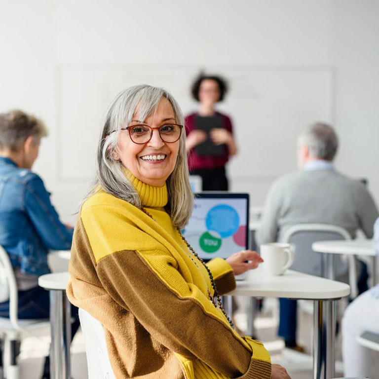 Smiling older woman in a classroom setting using a laptop, with other adults seated at tables and a presenter speaking in the background.